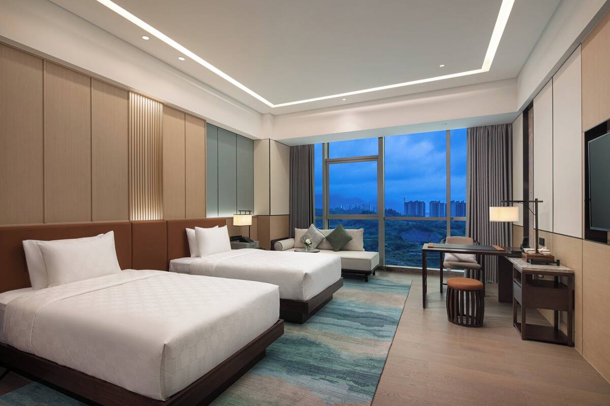 A hotel room with two beds, a desk, a chair, a TV, and a view of the cityscape.