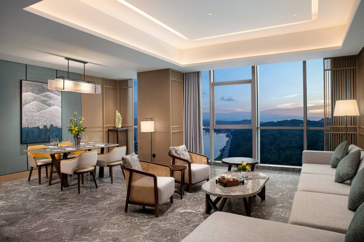A modern hotel room with a dining area, seating, and a large window offering a scenic view.