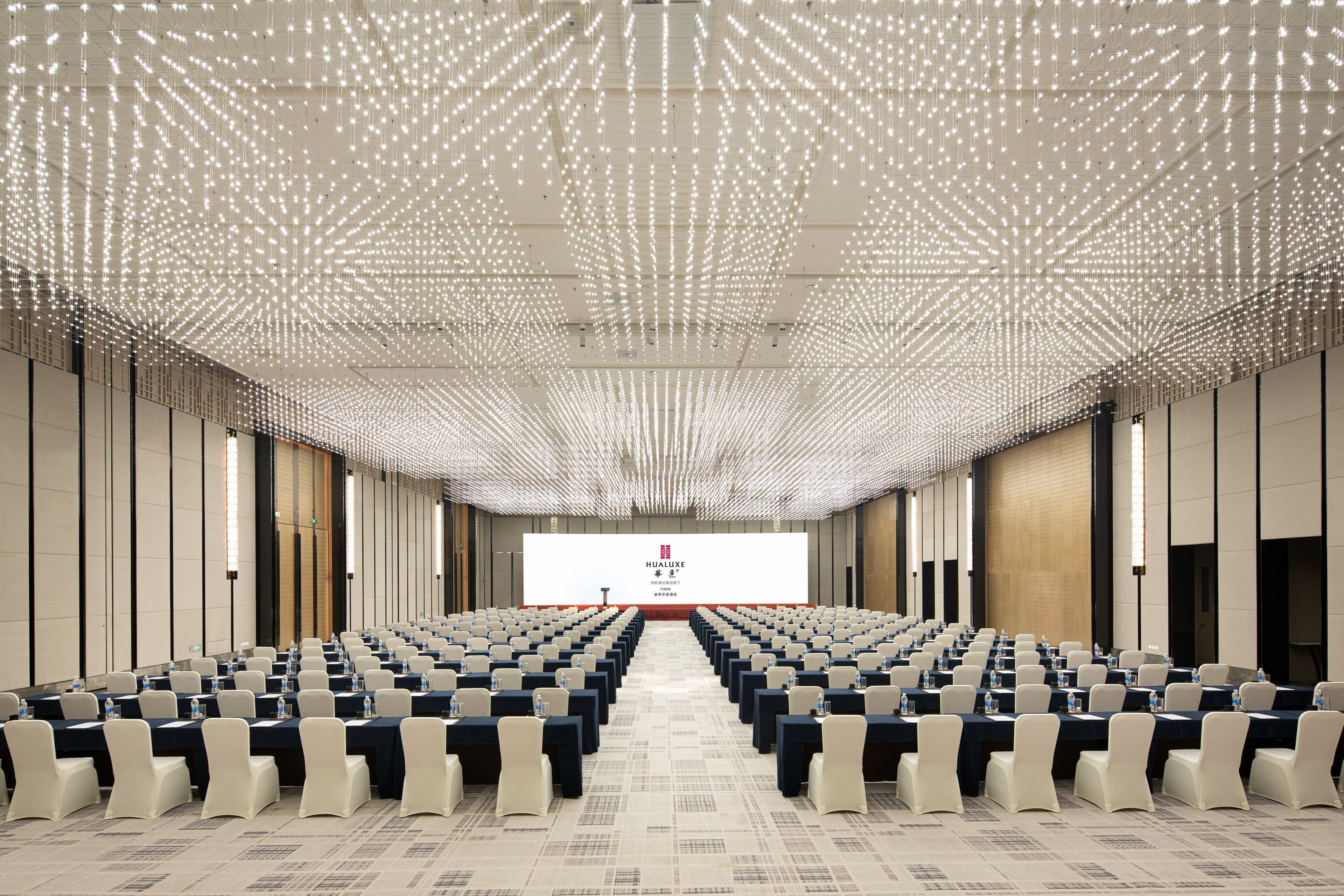A conference room with rows of tables and chairs, a large screen at the front, and a ceiling with a grid of lights.
