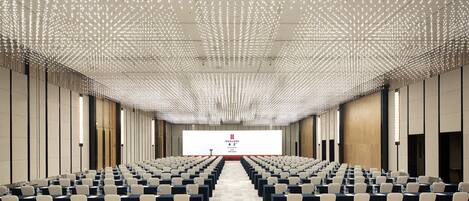 A conference room with rows of tables and chairs, a large screen at the front, and a ceiling with a grid of lights.