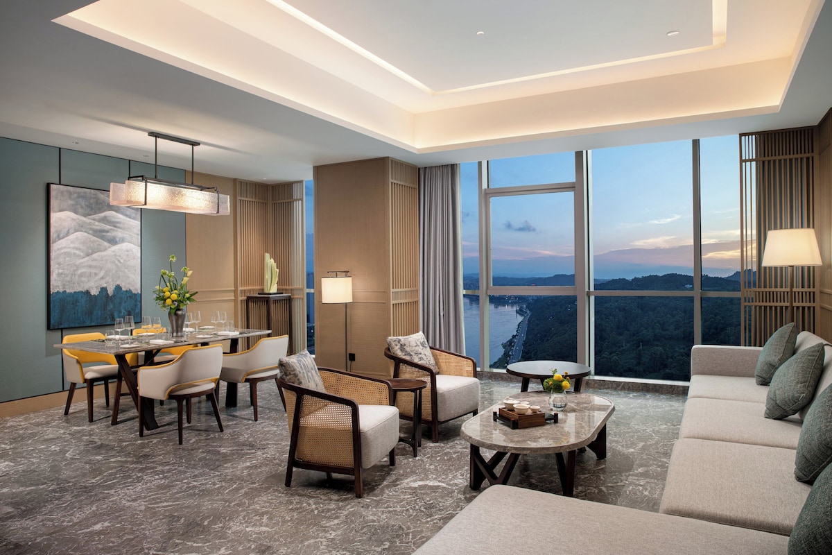 A modern hotel room with a dining area, seating, and a large window offering a scenic view.