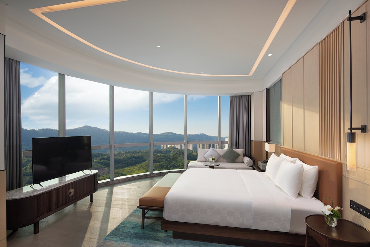A modern hotel room with a large bed, a flat-screen TV, and a view of a bridge and mountains.