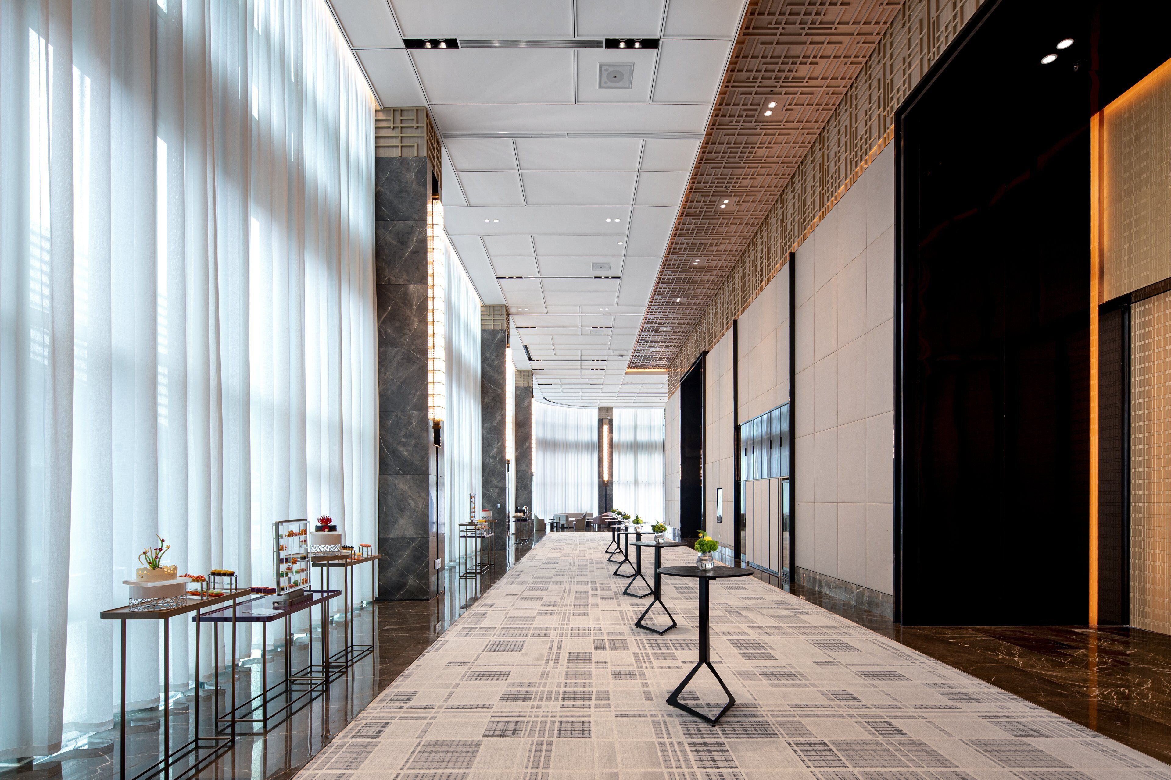 A modern hotel corridor with glass display tables, a patterned carpet, and a high ceiling with recessed lighting.