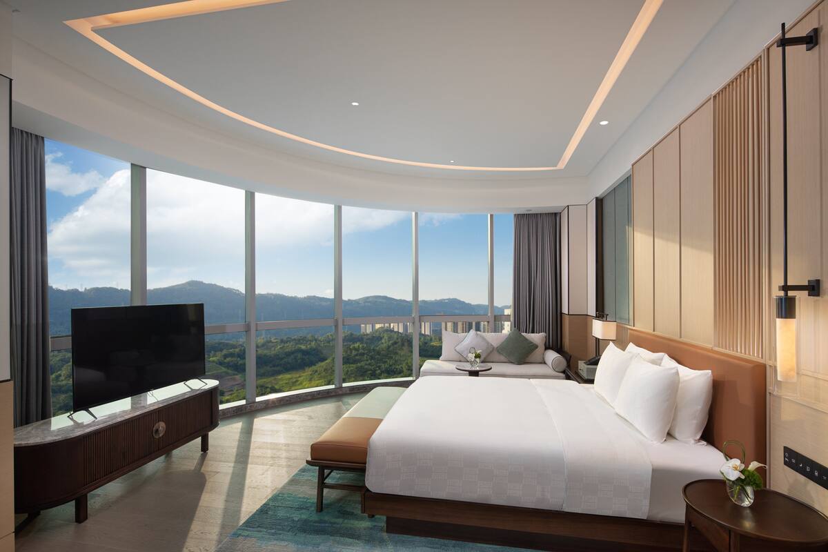 A modern hotel room with a large bed, a flat-screen TV, and a view of a bridge and mountains.
