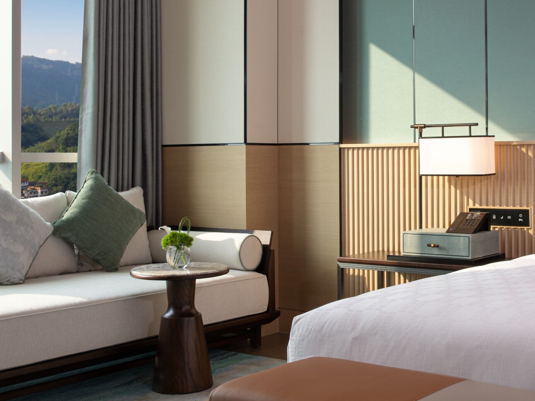 Habitación de hotel con cama, sofá, una mesita con un florero, lámpara y vistas al exterior.