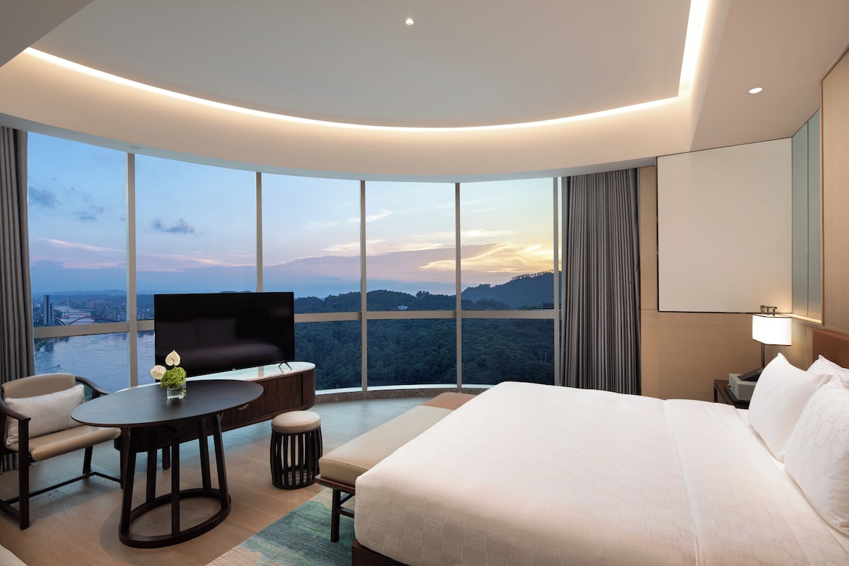 A modern hotel room with a large bed, a flat-screen TV, a dining table with chairs, and a view of a cityscape and mountains through large windows.