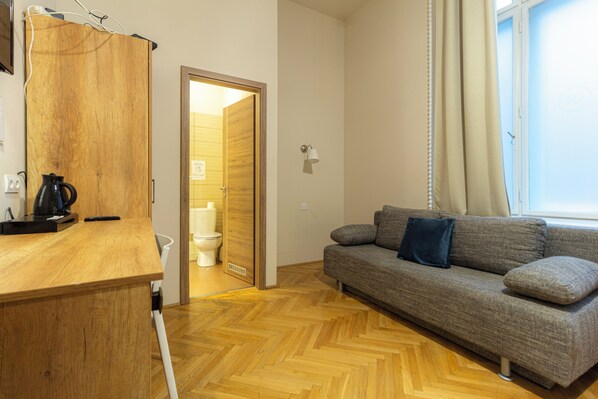 A room with a wooden desk, a grey sofa with cushions, a wooden floor, and a door leading to a bathroom.