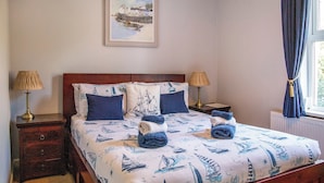 Una cama bien hecha con temática náutica, dos mesitas de noche con lámparas, un cuadro enmarcado sobre la cama y una ventana con cortinas.
