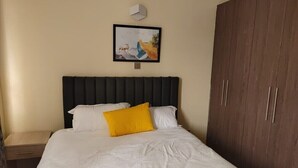 Un lit bien fait, avec des draps blancs et un seul oreiller jaune, une armoire en bois et un tableau encadré accroché au mur.