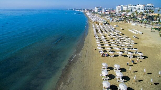 A beach with rows of sunbeds and umbrellas, a clear blue sea, and buildings in the background. - Les Palmiers Boutique Beach Hotel and Luxury Apartments in Larnaca (Cyprus, Larnaca)