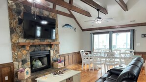 A living room with a stone fireplace, a flat-screen TV, a white coffee table, a black leather sofa, and a dining area with white chairs and a table. - Anchor's Away- Canal House with Private Dock (Holden Beach)