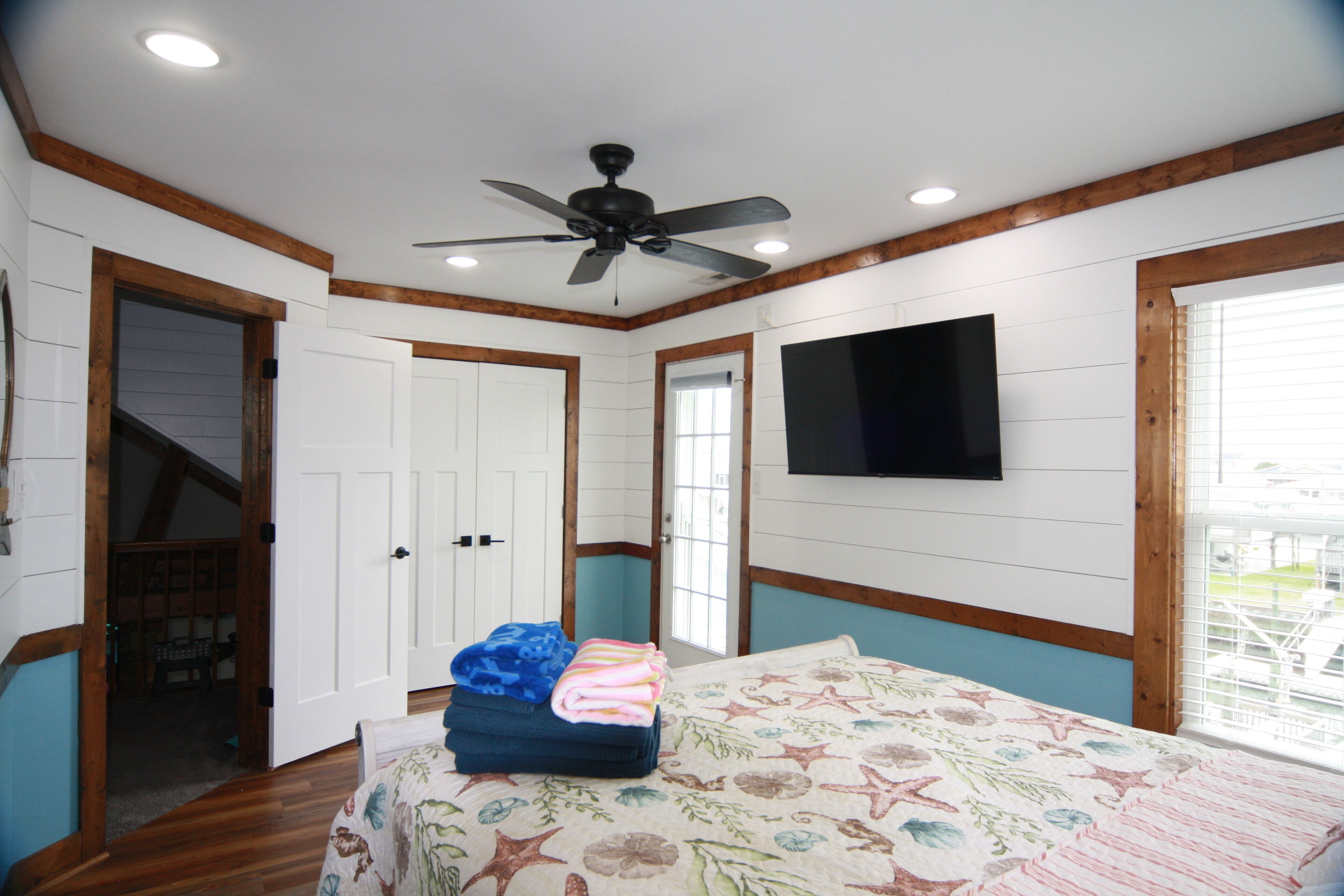 A bedroom with a bed, a ceiling fan, a TV mounted on the wall, and a window with blinds.