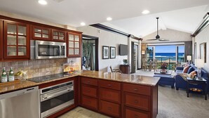 Cocina moderna con encimeras de granito, electrodomésticos de acero inoxidable y armarios de madera. Hay un comedor con vista al océano.