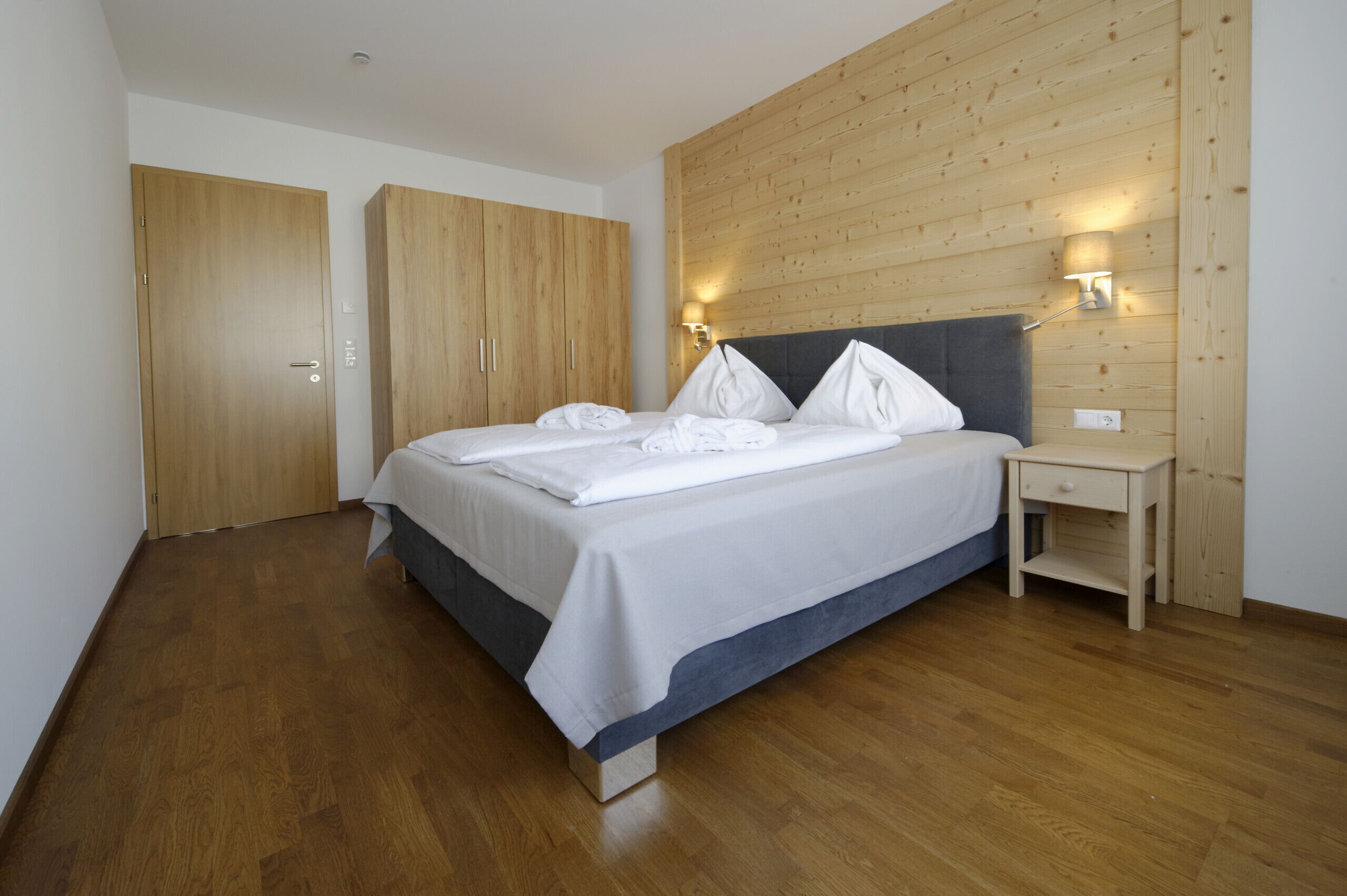 Un dormitorio con suelo de madera, cama con ropa blanca, armario de madera y mesita de noche con lámpara.