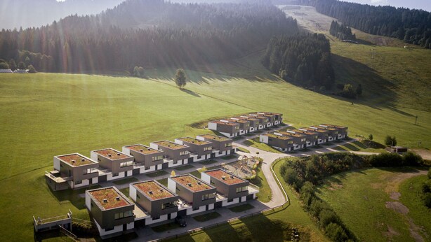 A modern residential complex with multiple buildings, green roofs, and a surrounding grassy area with hills in the background.