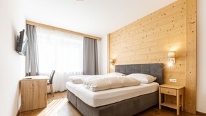 A modern bedroom with a wooden headboard, a bed with white linens, a desk with a chair, and a window with curtains.