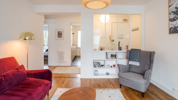 Una sala de estar moderna con un sofá rojo, un sillón gris y una mesa de centro de madera. Se observa una estantería con diversos objetos y un espejo grande en la pared.