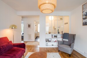 Un salon moderne avec un canapé rouge, un fauteuil gris et une table basse en bois. On y trouve une bibliothèque remplie de divers objets et un grand miroir accroché au mur.