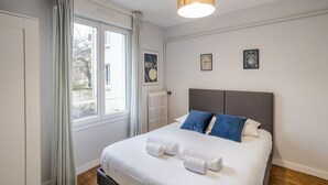 Una cama bien hecha con sábanas blancas y almohadas azules, una ventana con cortinas y un radiador.