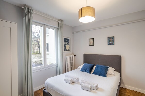 A neatly made bed with white linens and blue pillows, a window with curtains, and a radiator. - LE SÉVIGNÉ - Appartement cocooning à proximité du Thabor (Rennes)
