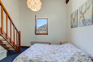 Un dormitorio con cama, ventana con vista a un bosque, una escalera de madera y dos cuadros colgados en la pared.