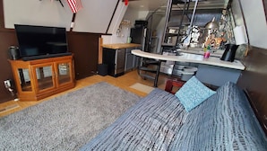 A compact living space with a TV on a wooden cabinet, a sofa with patterned cushions, a kitchen area with a counter and appliances, and a staircase leading to an upper level. - Table Rock Lake Cabin @ Black Oak Resort (Lampe)