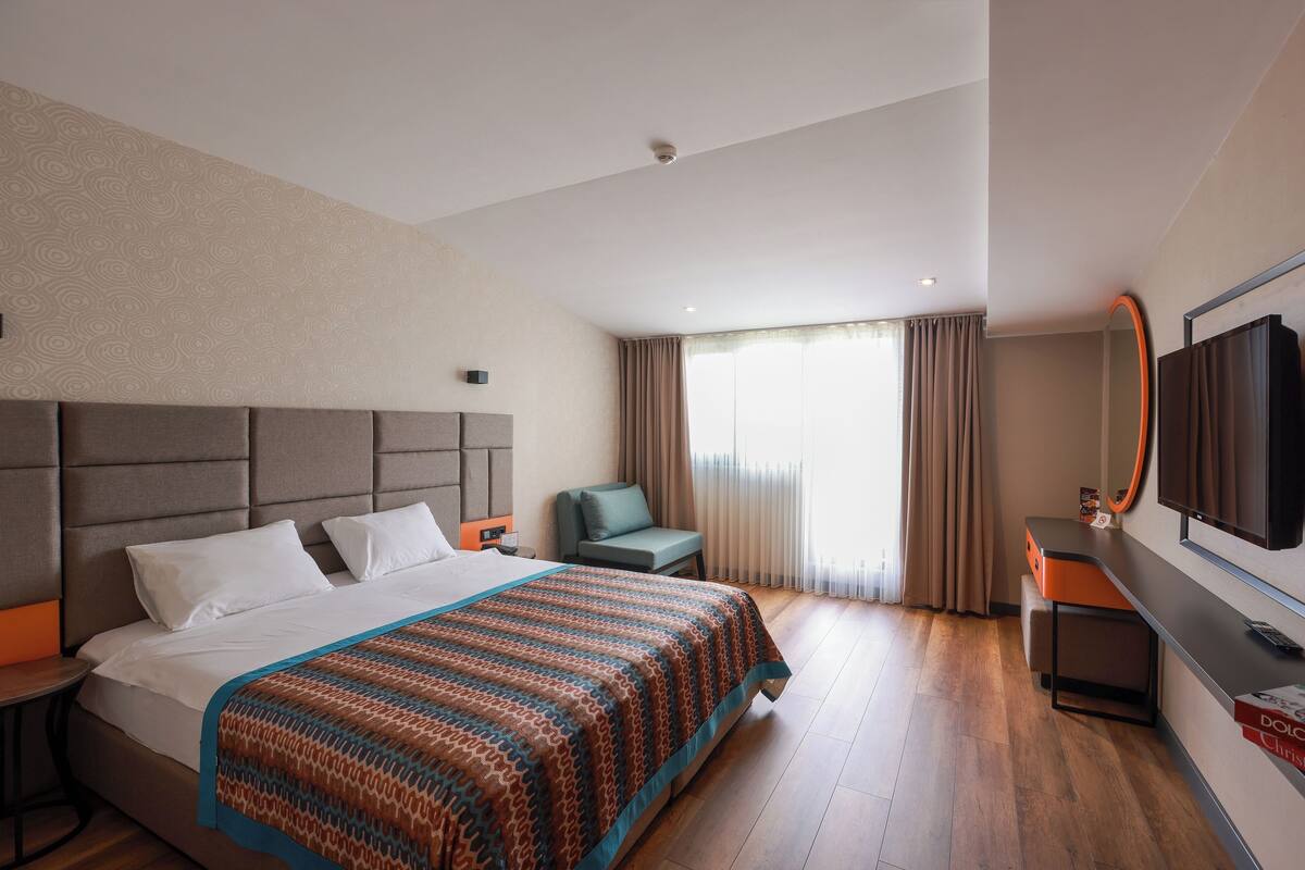 A hotel room with a large bed, a desk, a chair, a TV, and a window with curtains.