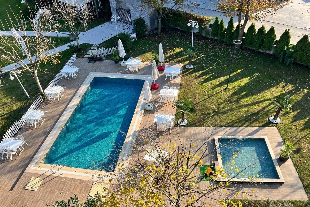 A rectangular swimming pool with a smaller adjacent pool, surrounded by white lounge chairs and umbrellas, set in a landscaped garden with a white picket fence and trees.