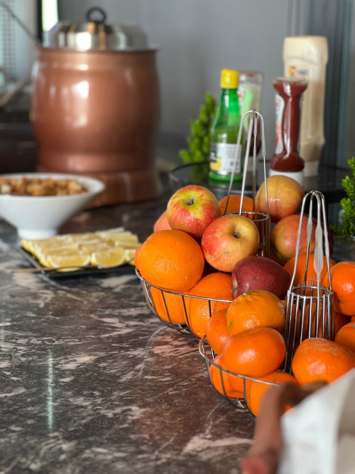 Uma bancada de cozinha com uma cesta de frutas contendo laranjas e maçãs, uma chaleira e temperos.