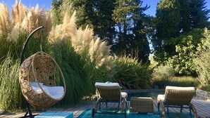 Una terraza con sillas de mimbre y una hamaca colgante, rodeada de árboles altos y pastos del pampa.