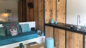 Un espacio de estar moderno con una pared de madera, una zona de cocina con fregadero y utensilios, y una acogedora sala de estar con cojines y un libro.