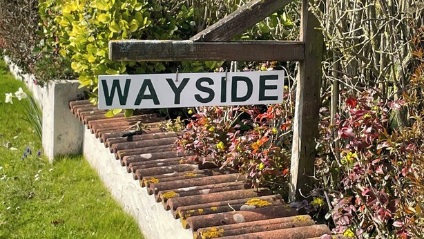 Naamplaatje met 'WAYSIDE' bevestigd aan een houten paal, naast een trap omringd door weelderige beplanting en bloeiende planten.