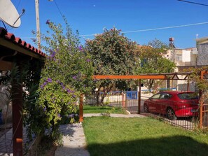 Un barrio residencial con un coche rojo estacionado en un camino de entrada cercado, un jardín con plantas en flor y un cielo azul despejado.