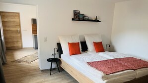 Een slaapkamer met een houten bed, wit beddengoed en oranje kussens. Er is een klein nachtkastje met een lamp en een wandplank boven het bed.
