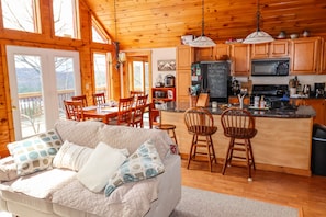 Interior acollidor d'una cabana, amb zona de menjador, cuina i sofà amb coixins estampats.