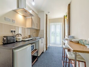 A compact kitchen with a toaster, kettle, microwave, and a small refrigerator. There is a window with blinds, a sink, and a counter with dishes.