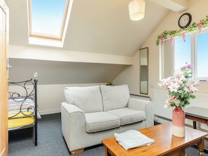 A cozy room with a grey sofa, a wooden table, a small window with a view, a clock on the wall, and a vase with flowers.