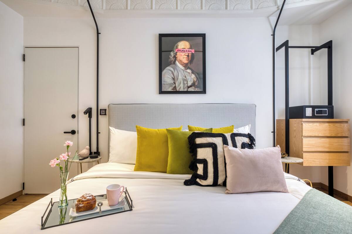 Camera d'albergo ordinata con un letto, cuscini, un comodino con un vassoio per la colazione, un'opera d'arte incorniciata al muro e una moderna lampada da comodino.