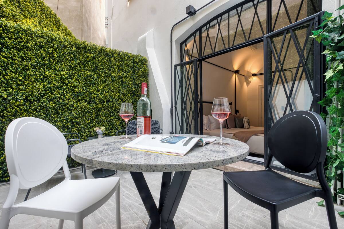 Un tavolo rotondo con due sedie, un libro e due bicchieri da vino su un patio con un siepe e un edificio sullo sfondo.