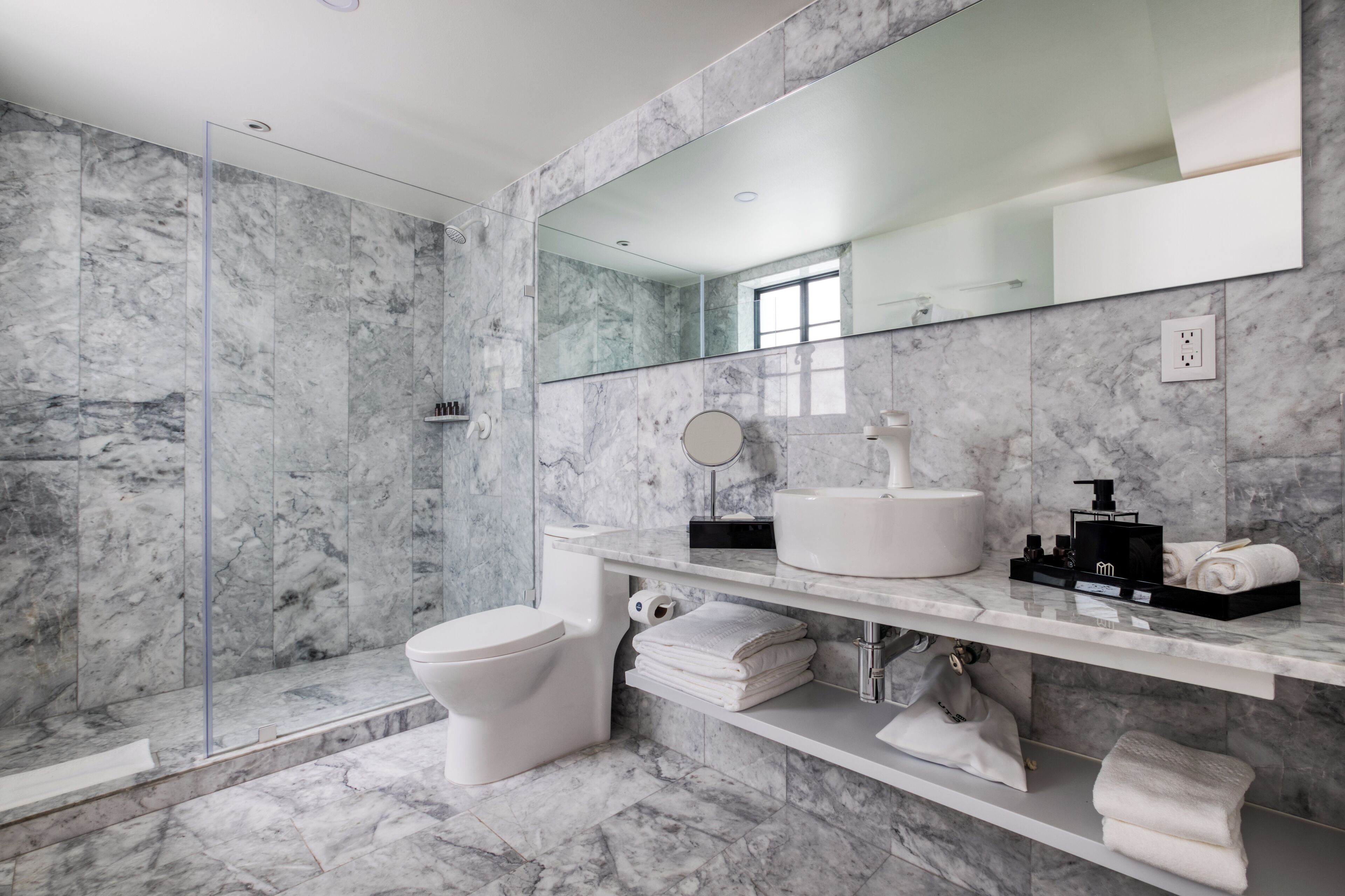 Een moderne badkamer met een marmeren vloer, een glazen douchecabine, een wit toilet, een wastafel met een zwarte zeepdispenser en een spiegel boven de wastafel.