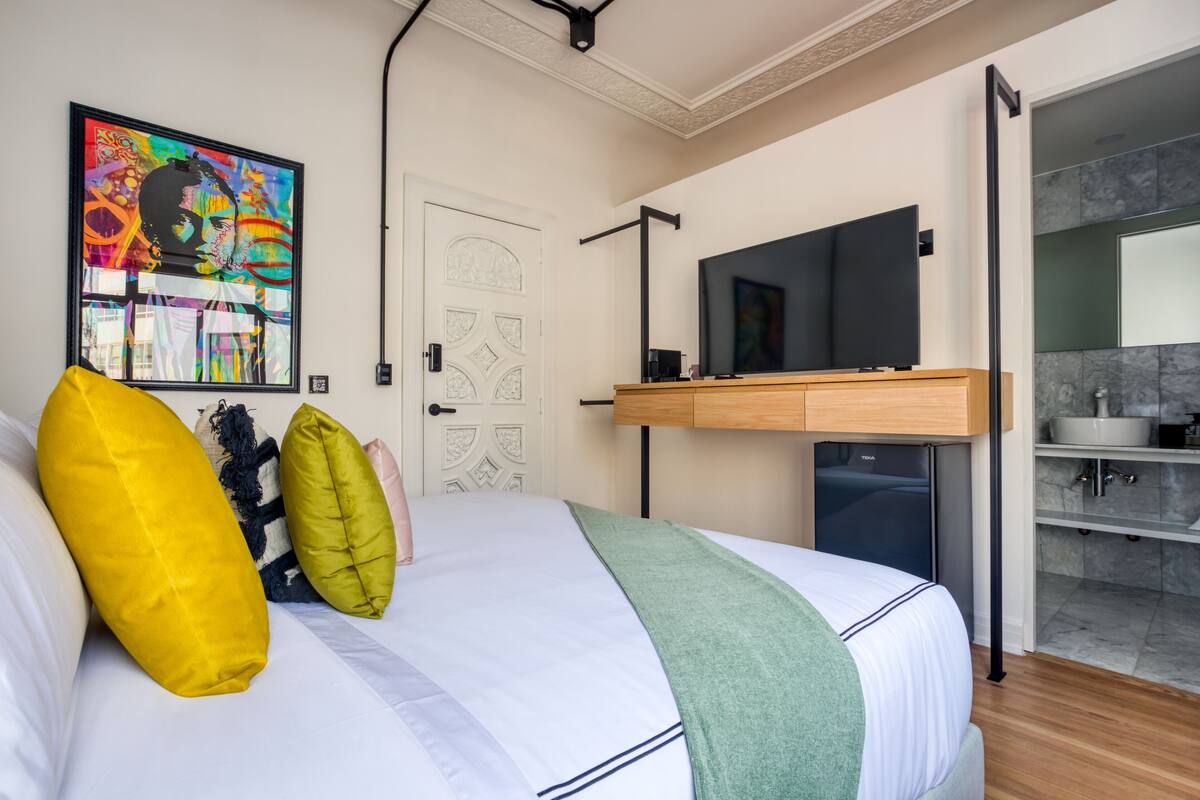 Una camera da letto moderna con un letto, un quadro di grandi dimensioni, una TV e un bagno visibile attraverso una porta aperta.