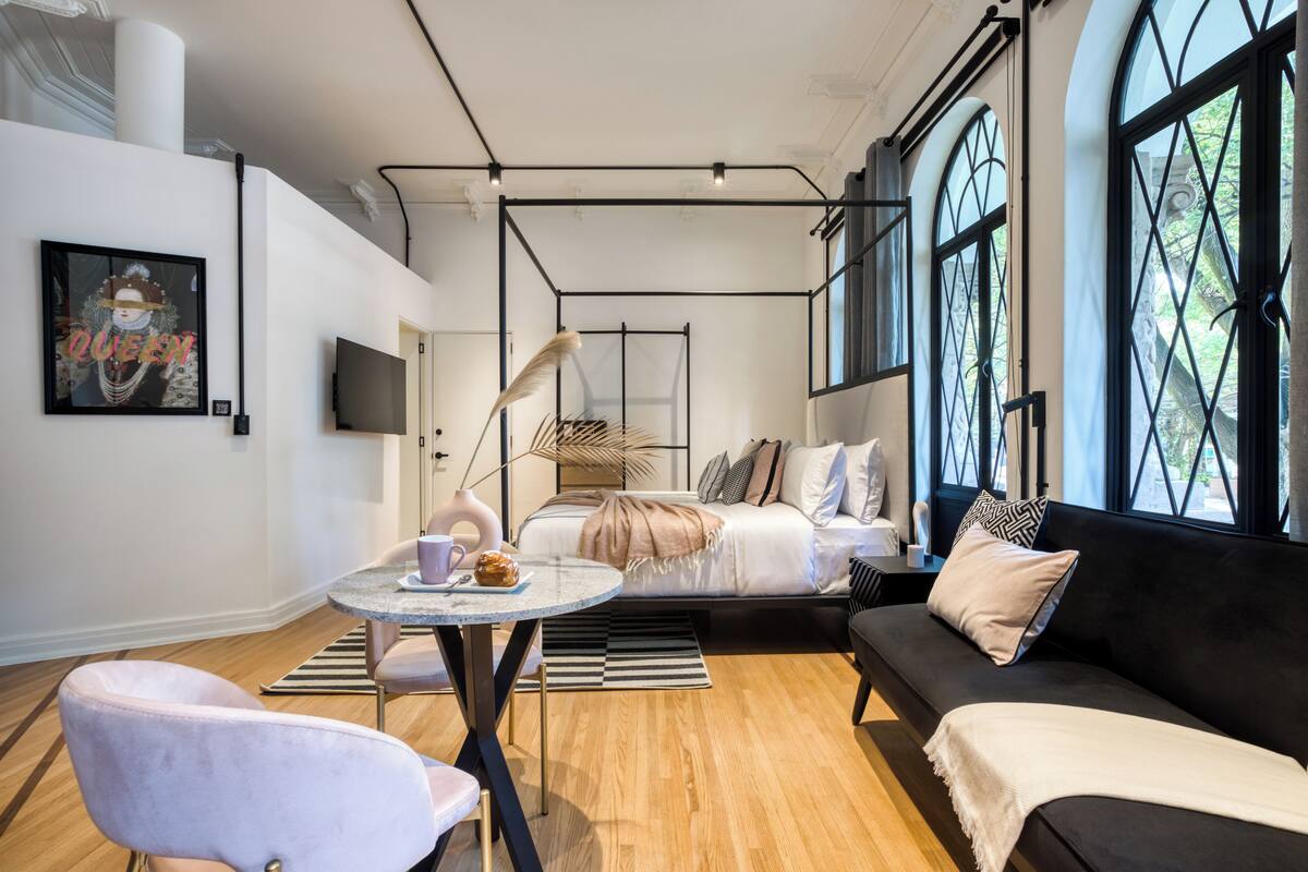 Un soggiorno moderno con divano, tavolo da pranzo con sedie, un letto con testiera e ampie finestre vista su vegetazione.