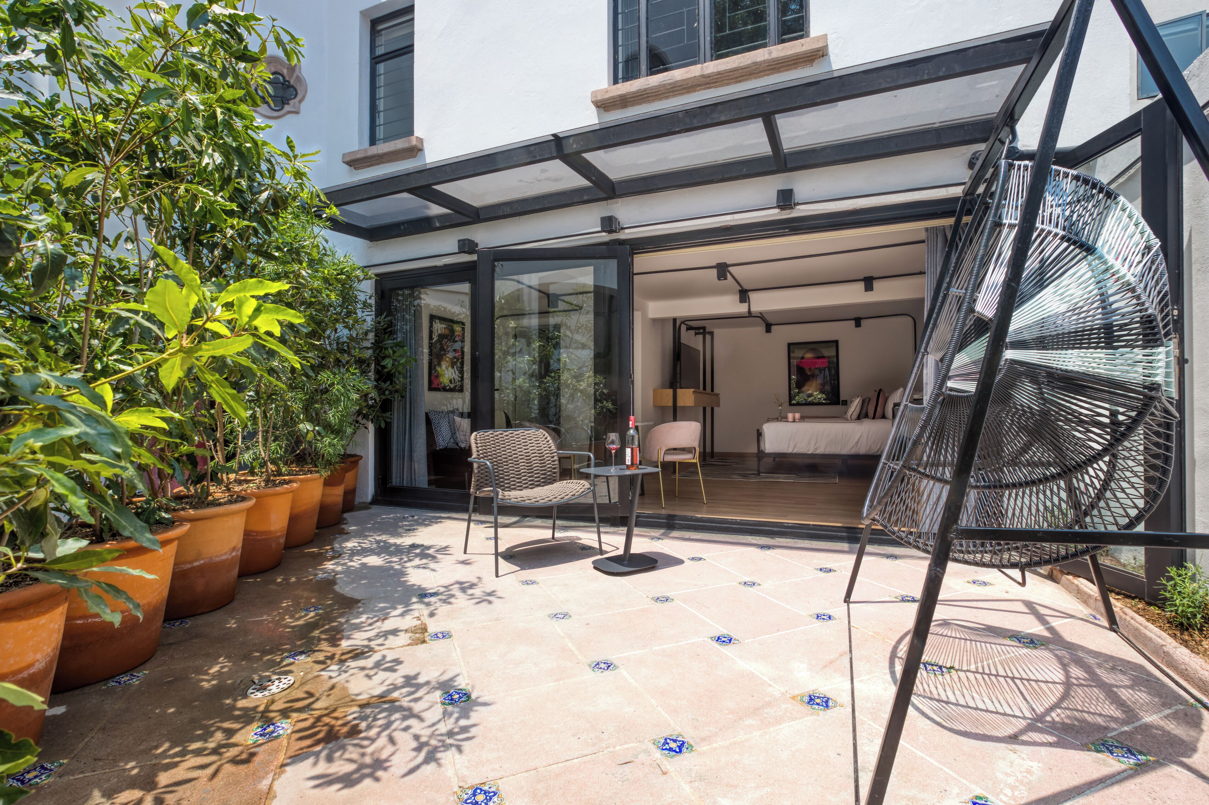 A modern patio with potted plants, a swing chair, and a glass-enclosed room with a bed and a desk.