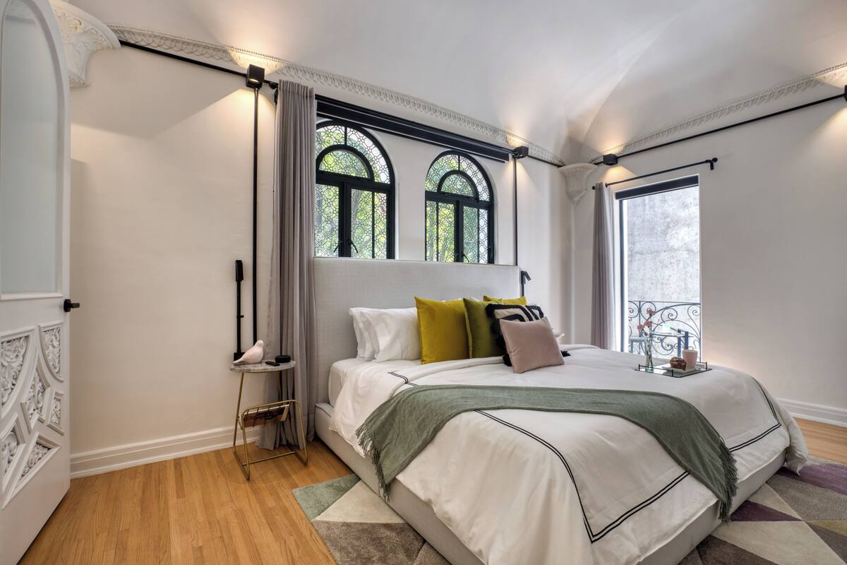 Camera da letto con un letto grande, due finestre con cornici nere, un tavolino con una sedia e un balcone con ringhiera.