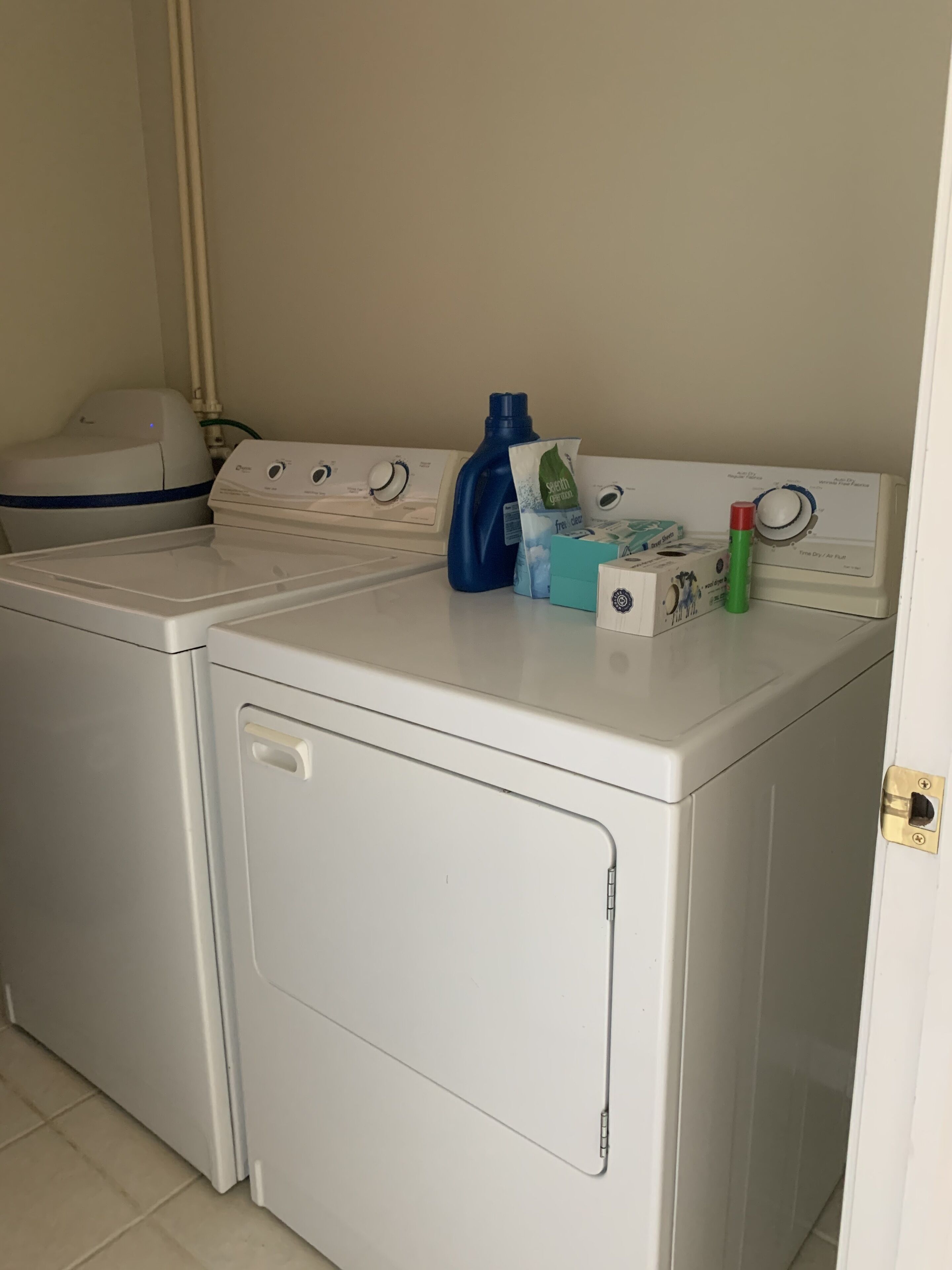Un espace dédié à la lessive, comprenant deux machines à laver superposées, une bouteille de lessive bleue, une bouteille verte et quelques dosettes de lessive.