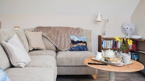A cozy living room with a beige sofa, a coffee table with a tray of cookies, a bookshelf, and a fan. - 1 bedroom accommodation in Great Bealings (Great Bealings)