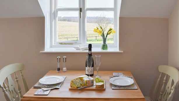 A dining table set for two with plates, cutlery, a bottle of wine, a glass, and a vase with flowers by a window. - 1 bedroom accommodation in Great Bealings (Great Bealings)