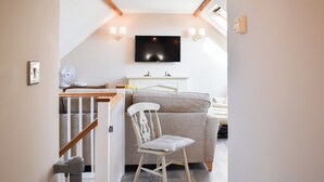 A cozy living room with a wooden staircase, a beige sofa, a white chair, and a flat-screen TV mounted on the wall. - 1 bedroom accommodation in Great Bealings (Great Bealings)