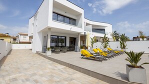 Una casa moderna de dos pisos con un patio que incluye sillas de descanso amarillas y plantas en macetas.
