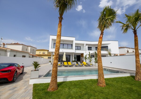A modern house with a swimming pool, palm trees, and a red sports car parked in front. - A magnificent contemporary villa right in the city center, a 3-minute walk from the beach and restaurants. (Empuriabrava)
